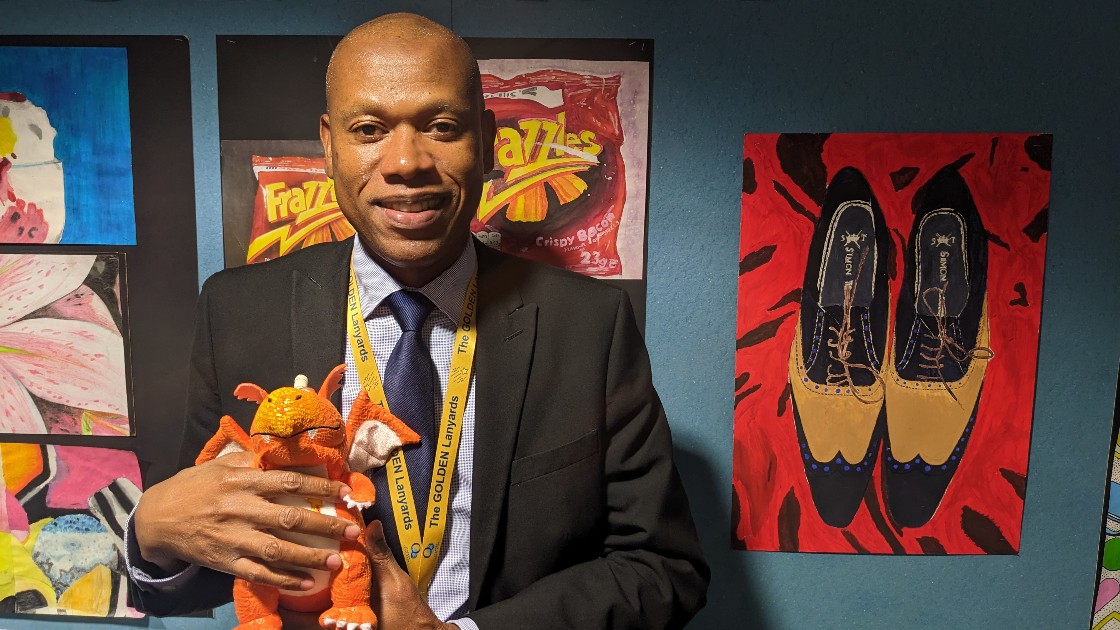Teacher holding a toy dragon in front of student artwork on a wall