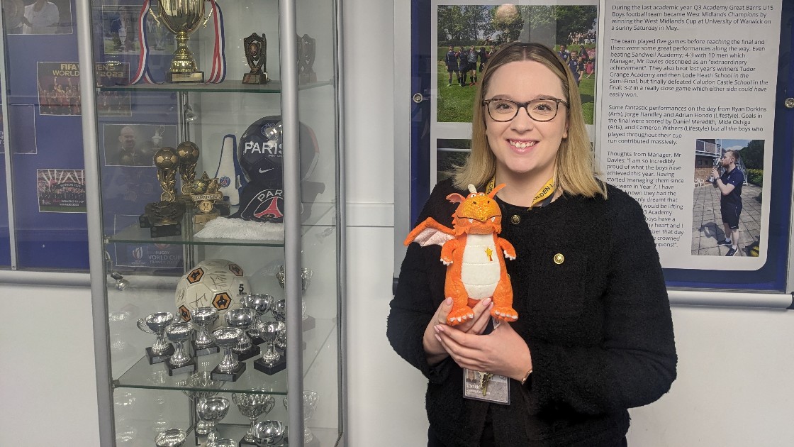 A teacher holding a dragon toy stands in front of a trophy display case